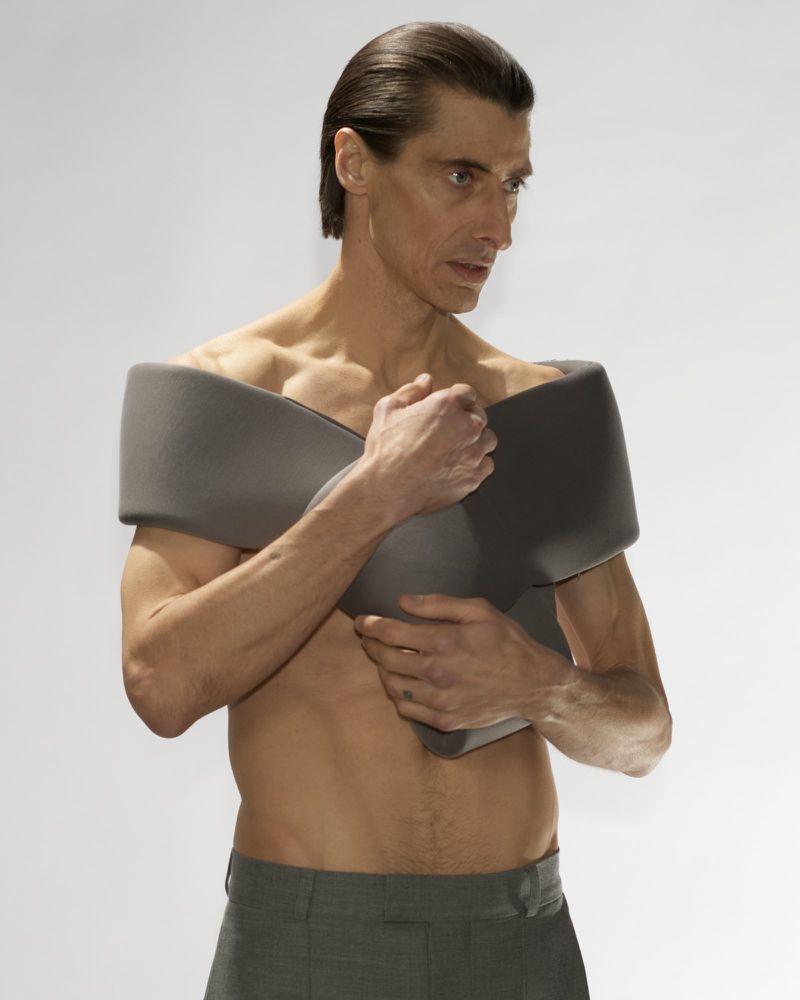 Adult male poses shirtless with gray fabric over his shoulders in a studio setting, looking away from the camera.