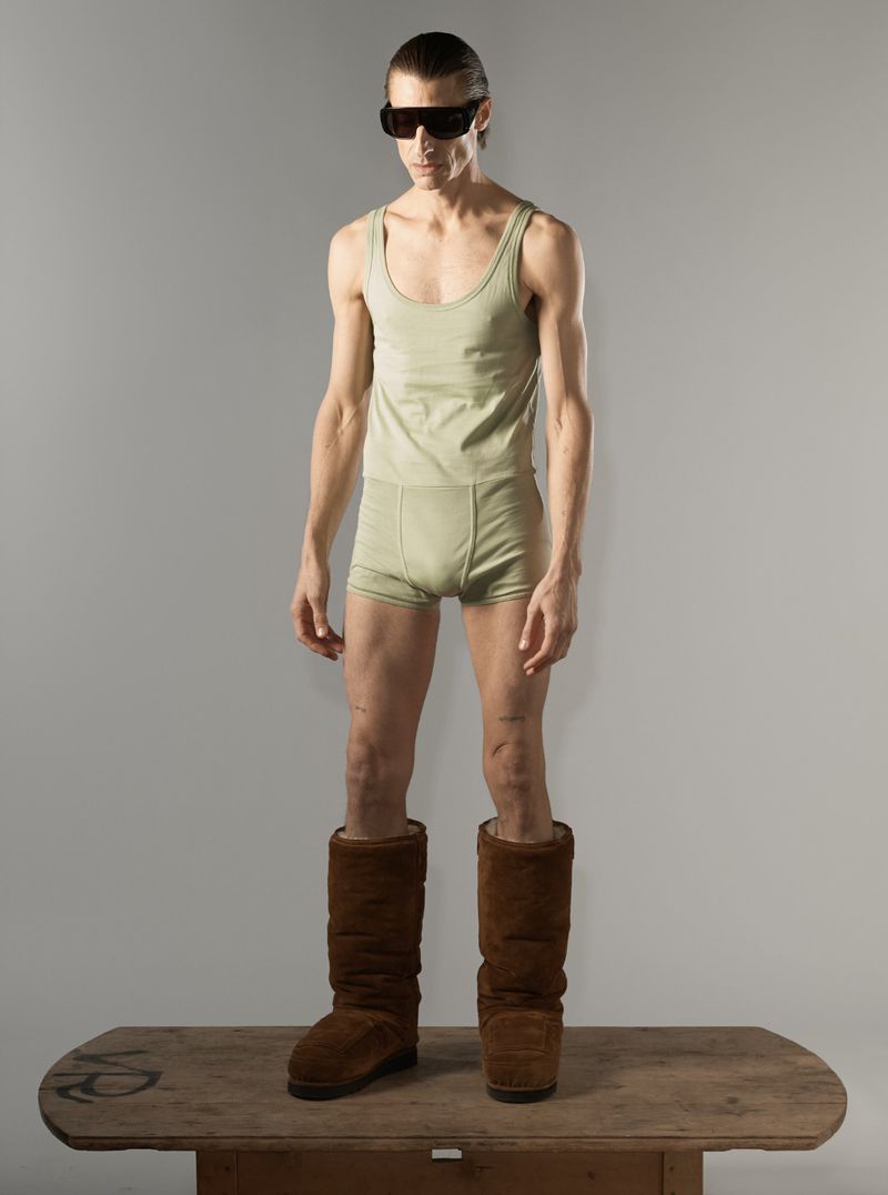 Adult male stands on a wooden table in a studio, wearing a tank top, shorts, sunglasses, and oversized boots against a gray background.