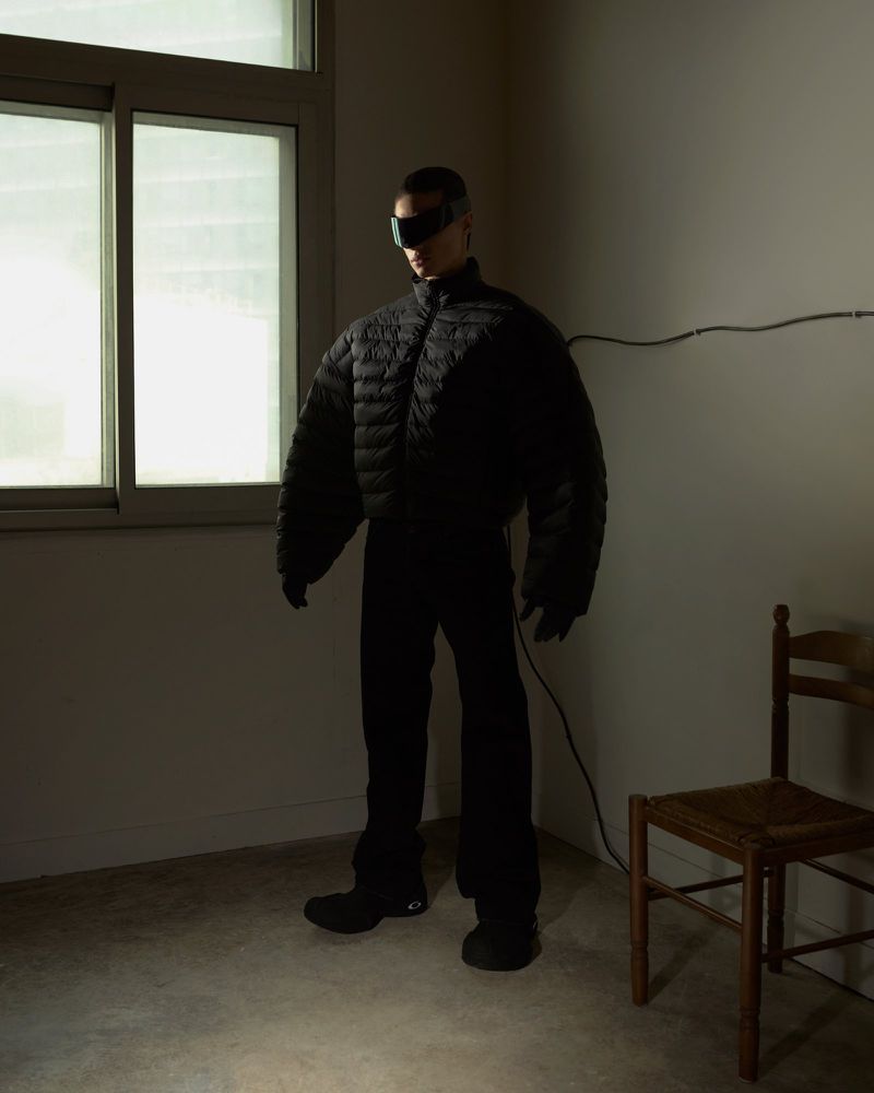 Man in black puffer jacket and futuristic visor stands indoors with natural window light and a wooden chair in dim corner.