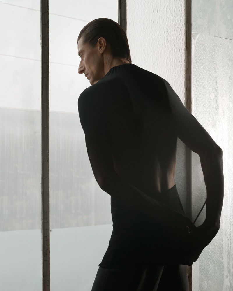 Adult man in black top and shorts stands by a window looking out; indoor setting, neutral colors.