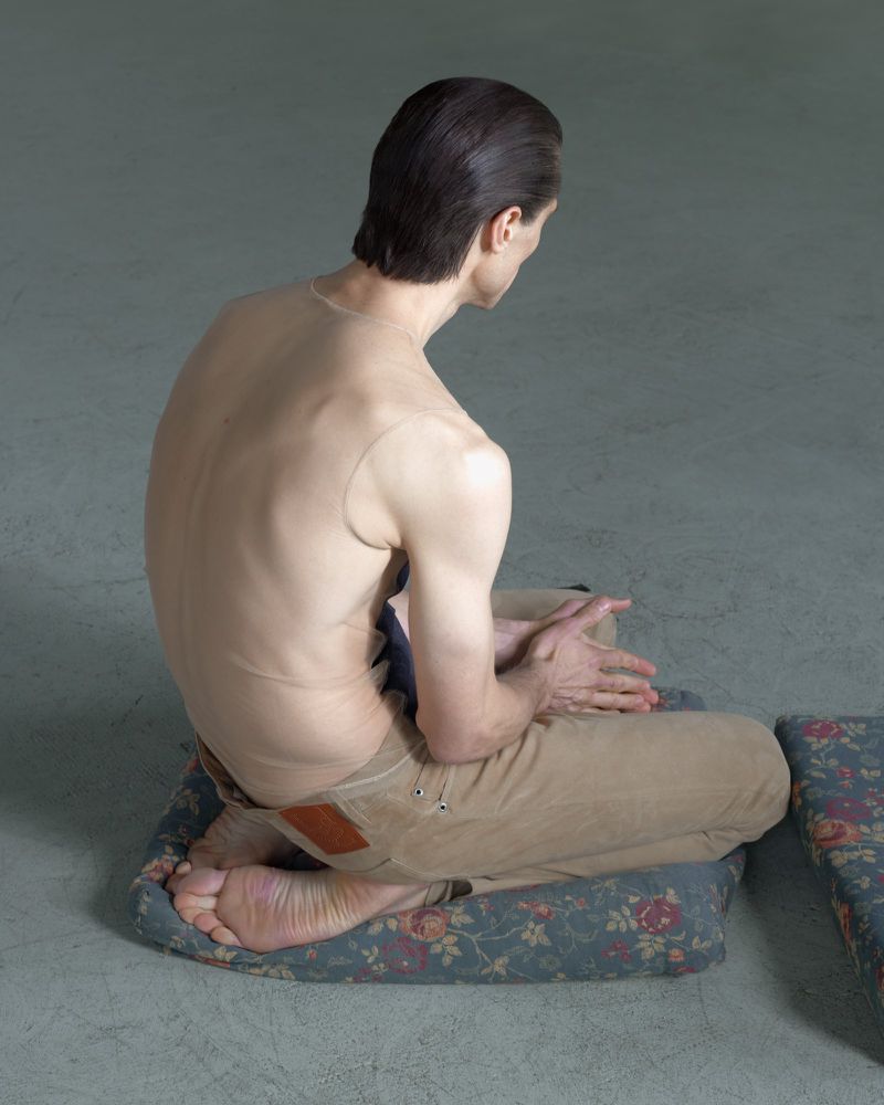A slender, light-skinned adult is seated cross-legged on a floral cushion, facing away, in an indoor studio with diffused lighting.