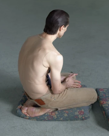 A slender, light-skinned adult is seated cross-legged on a floral cushion, facing away, in an indoor studio with diffused lighting.