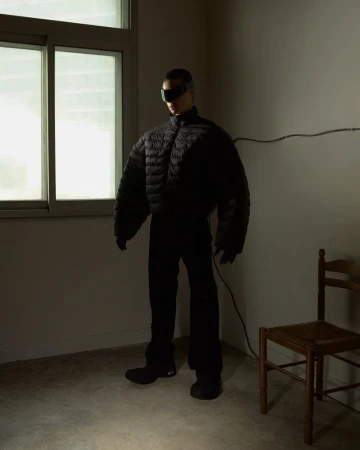 Model wearing a black puffer jacket and futuristic sunglasses standing indoors near a window and wooden chair