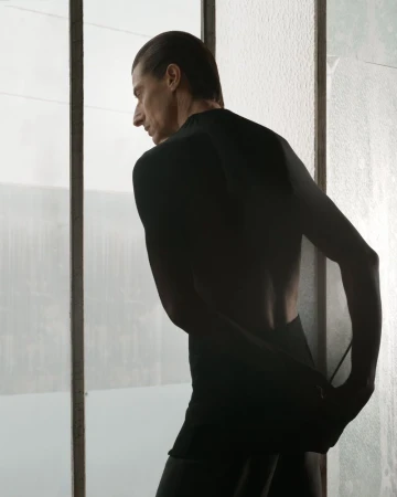 Male model in a black top standing in profile against a tall window with vertical frames