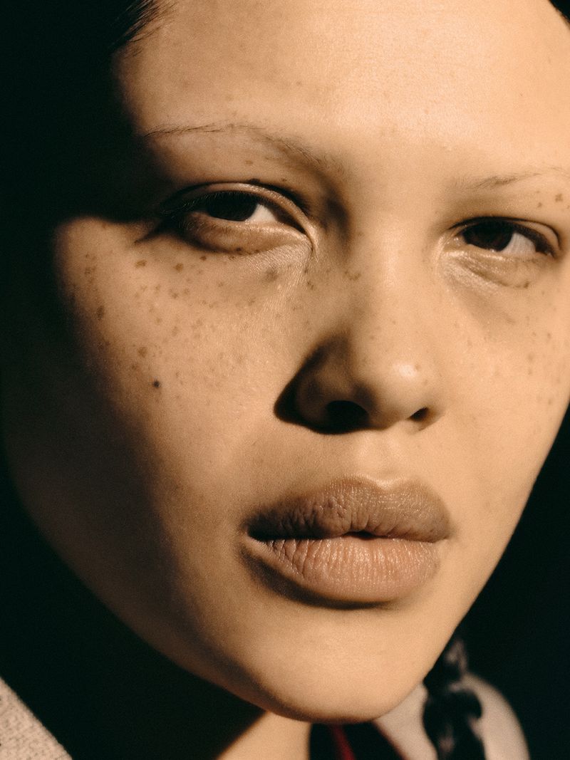 Close-up portrait of a woman with light skin, freckles, dark eyes, light eyebrows, and dark hair braided down the back of her head.