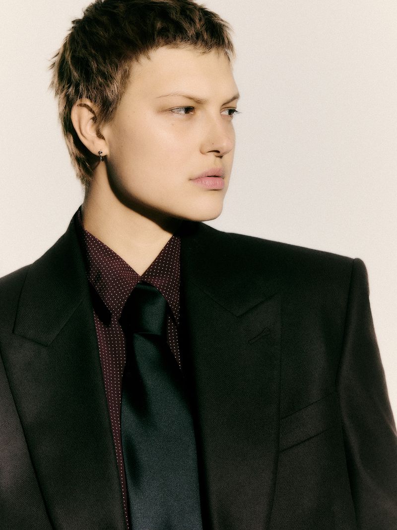 Woman in a dark suit, wearing a patterned collared shirt and tie, poses against a light background in a studio portrait.