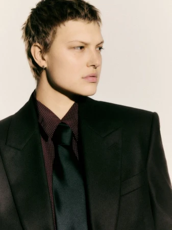 Woman in a dark suit, wearing a patterned collared shirt and tie, poses against a light background in a studio portrait.