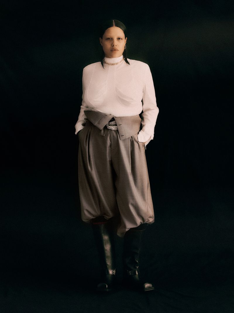 Fashion shot of a woman wearing an ecru-colored, long-sleeve blouse, high-waisted grey pants, and black boots against a dark background.