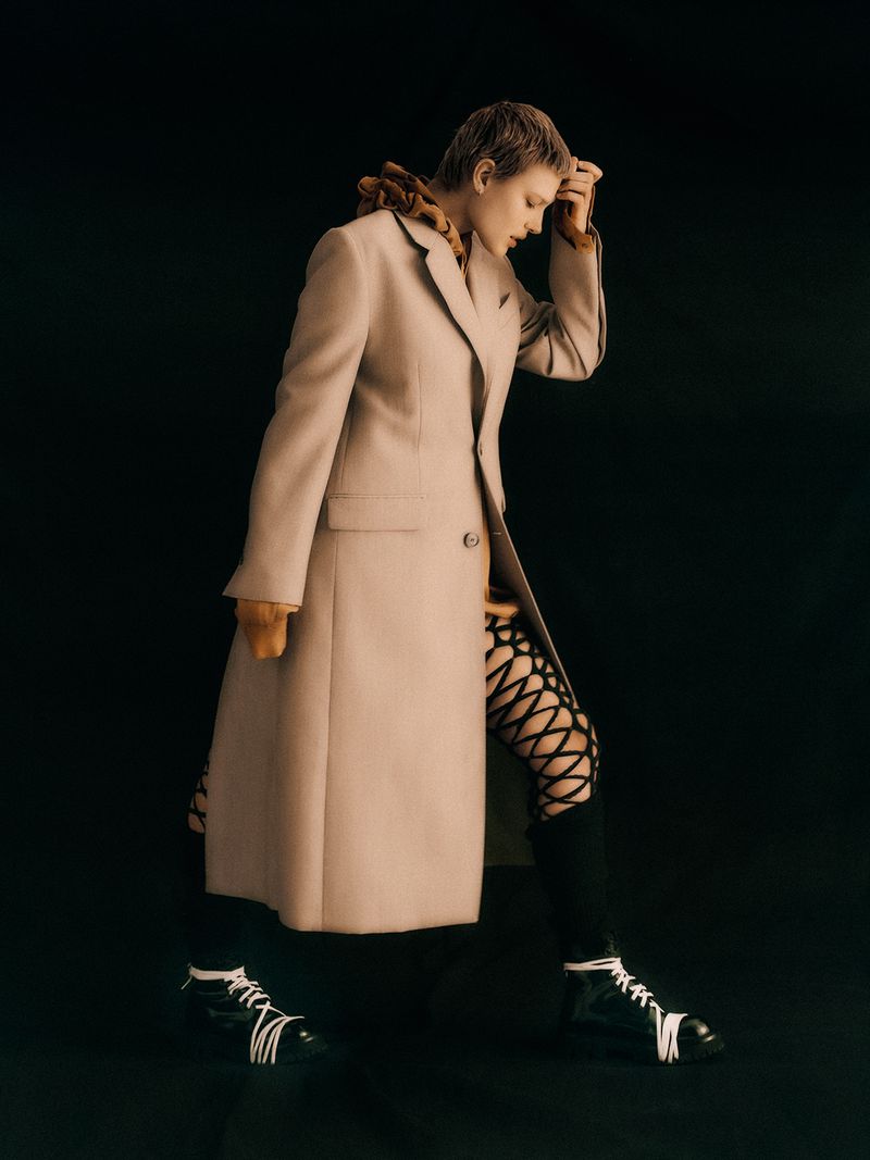 Fashionable woman in a tan coat and patterned tights walks against a dark backdrop. The shot highlights style and clothing.
