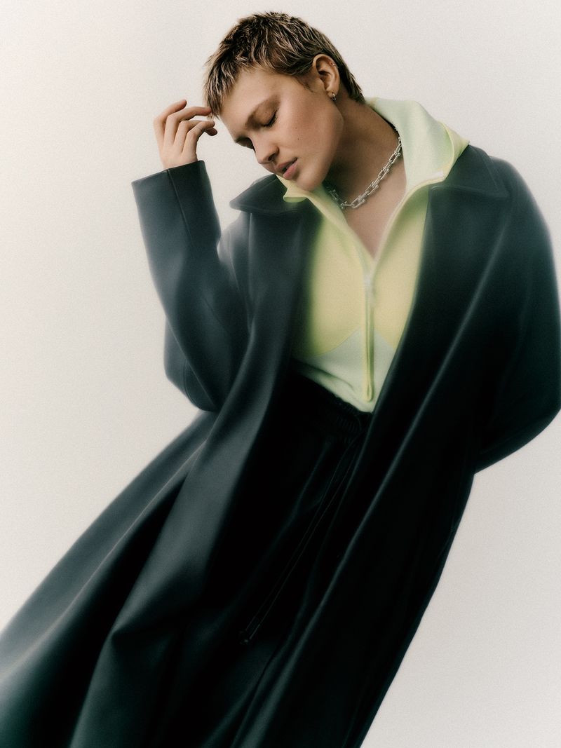 Adult posing in a dark coat, light-yellow top, and silver necklace in a studio with a soft neutral background.