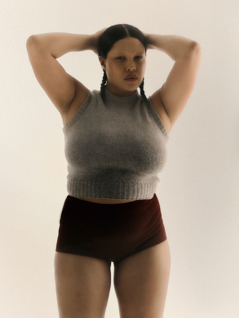 Portrait of a woman with braids, wearing a grey top and maroon shorts against a neutral background, posing with hands behind her head.