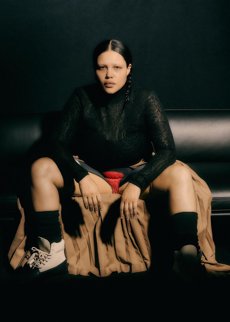 Woman in black top and brown skirt sitting indoors with black background. The woman wears white sneakers with black socks.