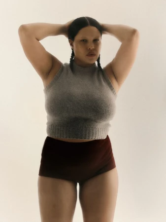Model with braided dark hair wearing a grey sleeveless knit top and dark shorts posing against a seamless white background.
