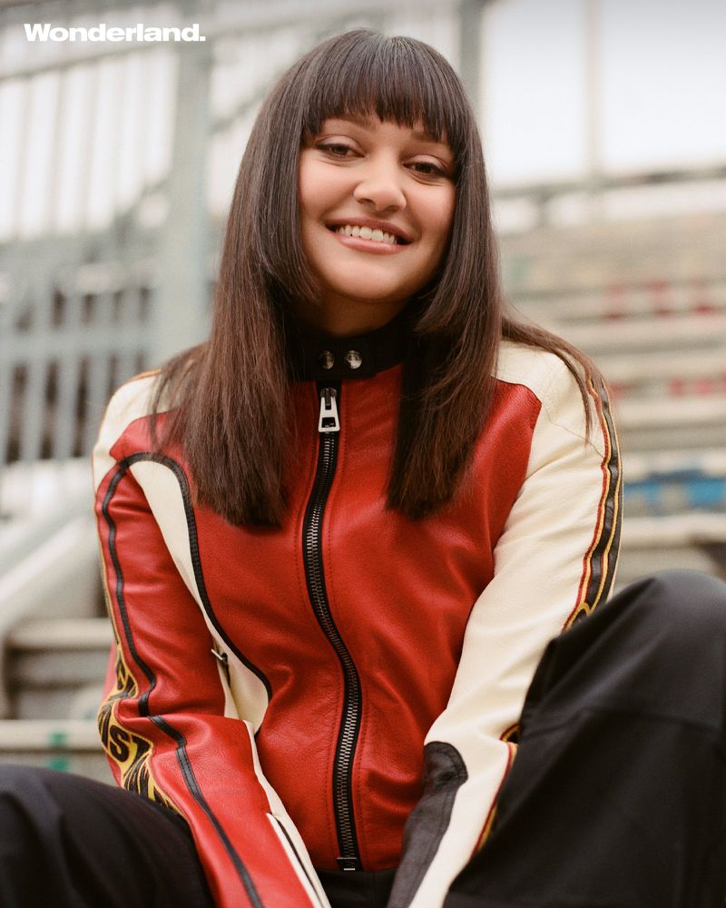 Smiling woman with bangs in red-and-white racing jacket poses in front of an industrial background in an eye-level medium shot.