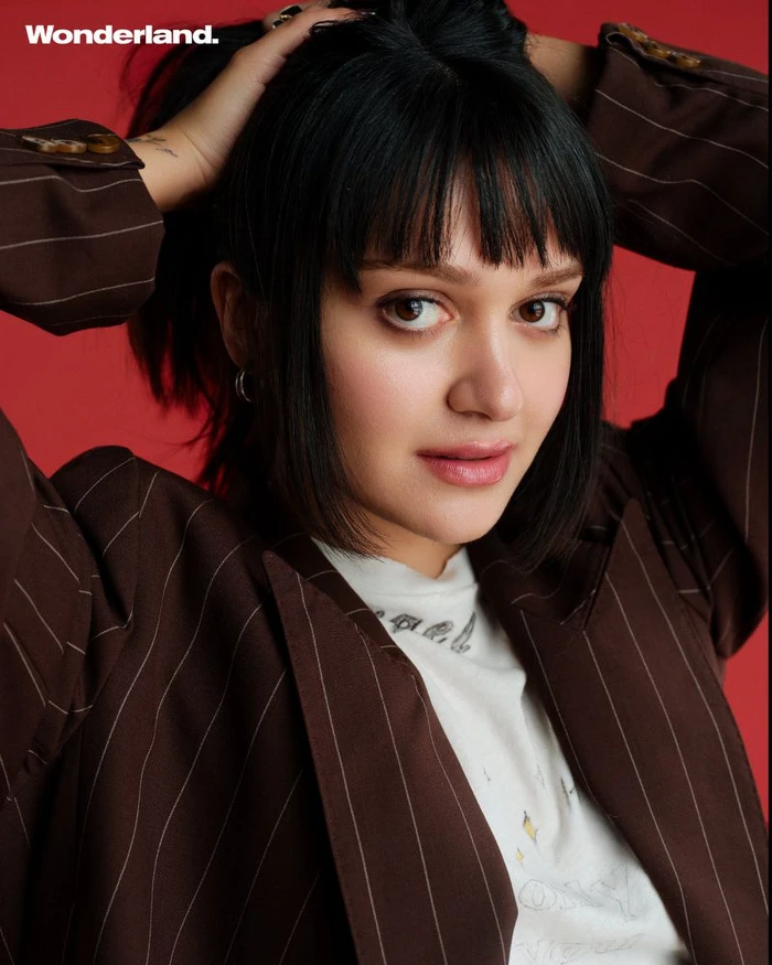 Ariela Barer wearing a brown pinstripe blazer against a solid red background for Wonderland Magazine, photographed by Tianzheng Yun.