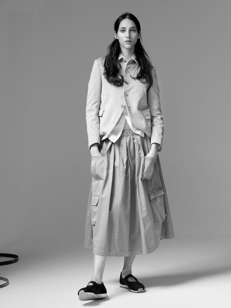 Monochrome fashion shot of a young adult woman in a light beige blazer, white top, and a beige pleated skirt in a studio setting.