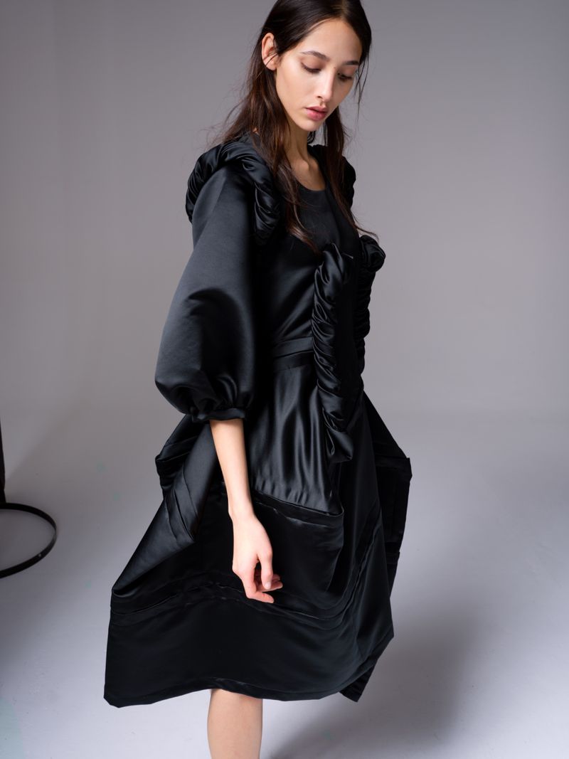 A young woman poses indoors wearing a voluminous black satin dress with puffed sleeves in a fashion setting with a grey background.