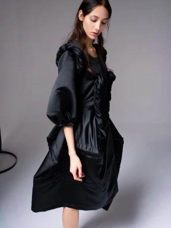 A young woman poses indoors wearing a voluminous black satin dress with puffed sleeves in a fashion setting with a grey background.