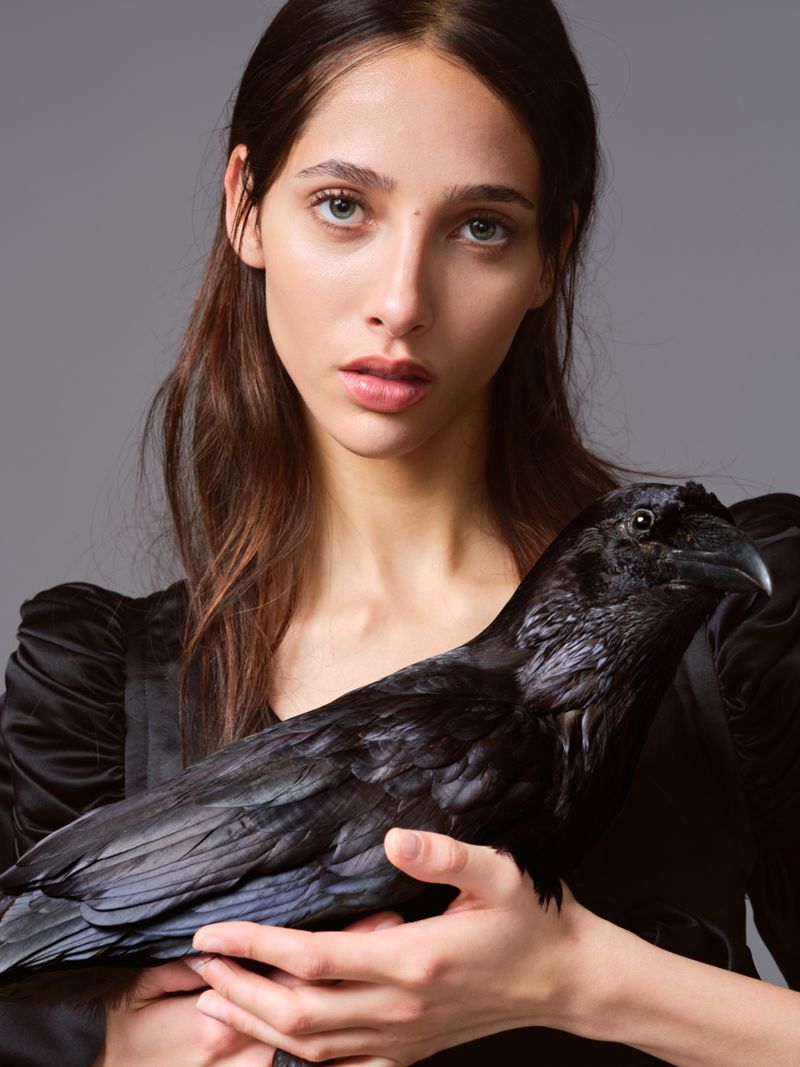 Close-up studio shot: Young woman holds large black raven. She wears a black dress. Grey background. Moody, neutral color grading.
