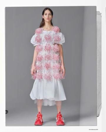 Model Yasmin Wijnaldum wearing a white dress adorned with pink floral clusters and red sneakers against a grey studio backdrop.