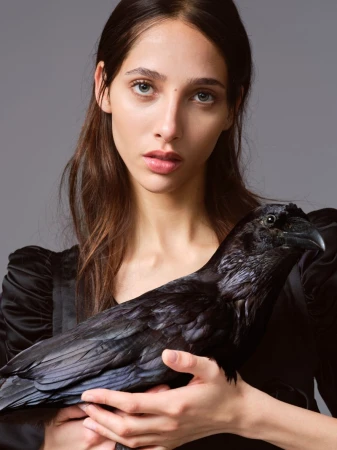 Model Yasmin Wijnaldum holding a black bird against a plain grey background in a portrait for At Large Magazine.