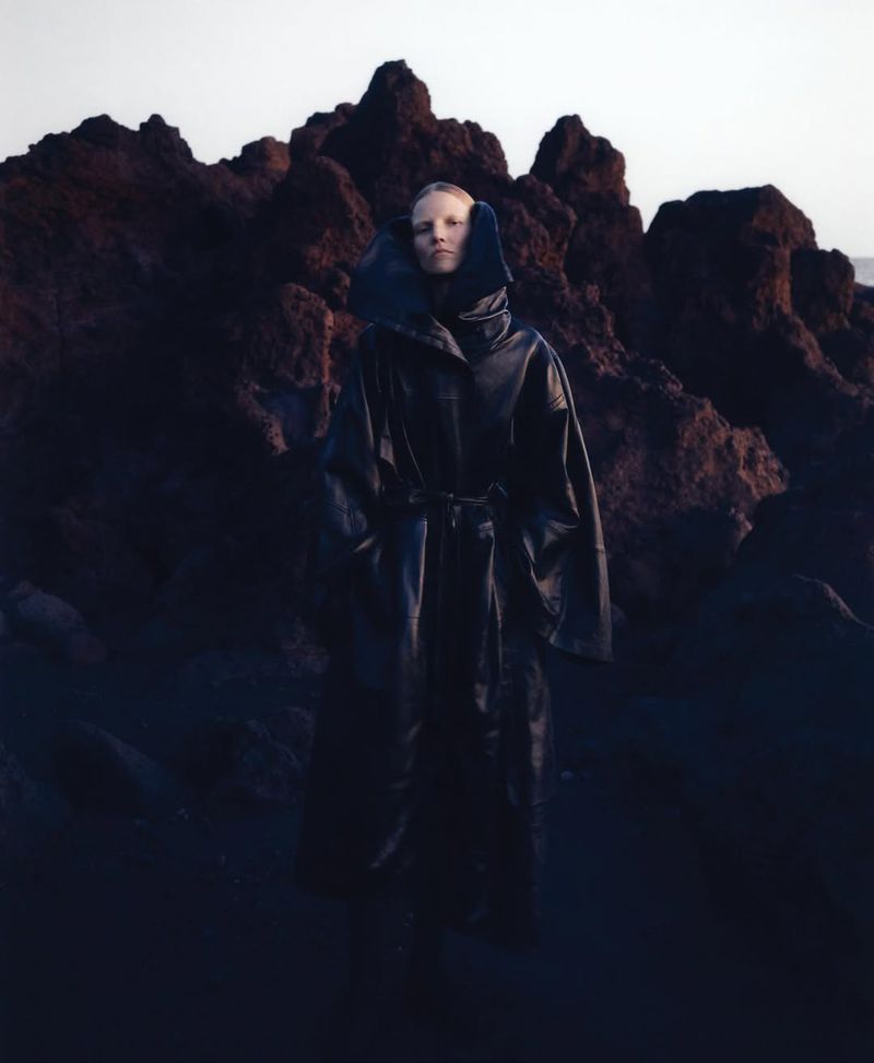 Adult female model in a full-length dark coat with a belt, standing outdoors in front of a dark rocky landscape and soft light.