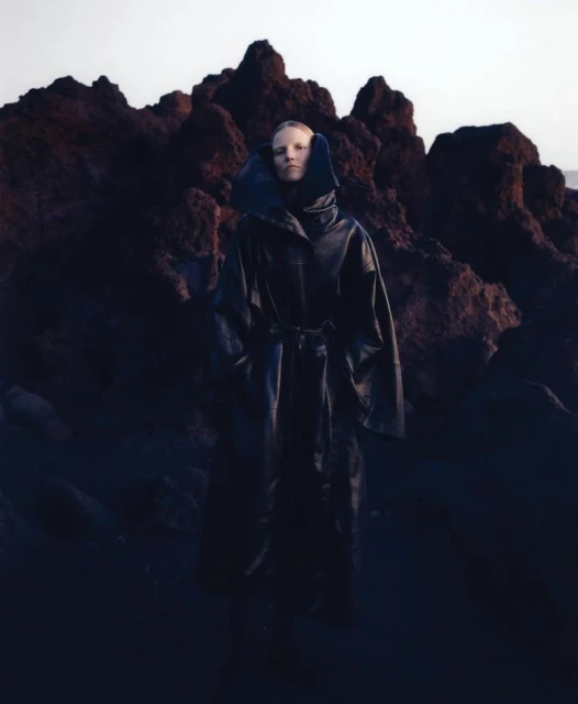 Adult female model in a full-length dark coat with a belt, standing outdoors in front of a dark rocky landscape and soft light.