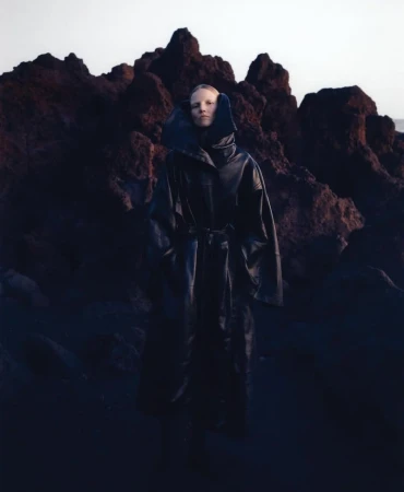 Adult female model in a full-length dark coat with a belt, standing outdoors in front of a dark rocky landscape and soft light.