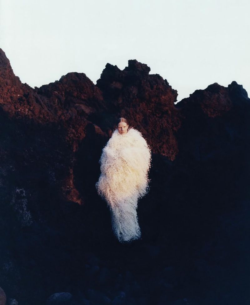 A woman with long hair stands amidst black volcanic rock wearing a long, fluffy, white garment against a light sky in a medium shot.
