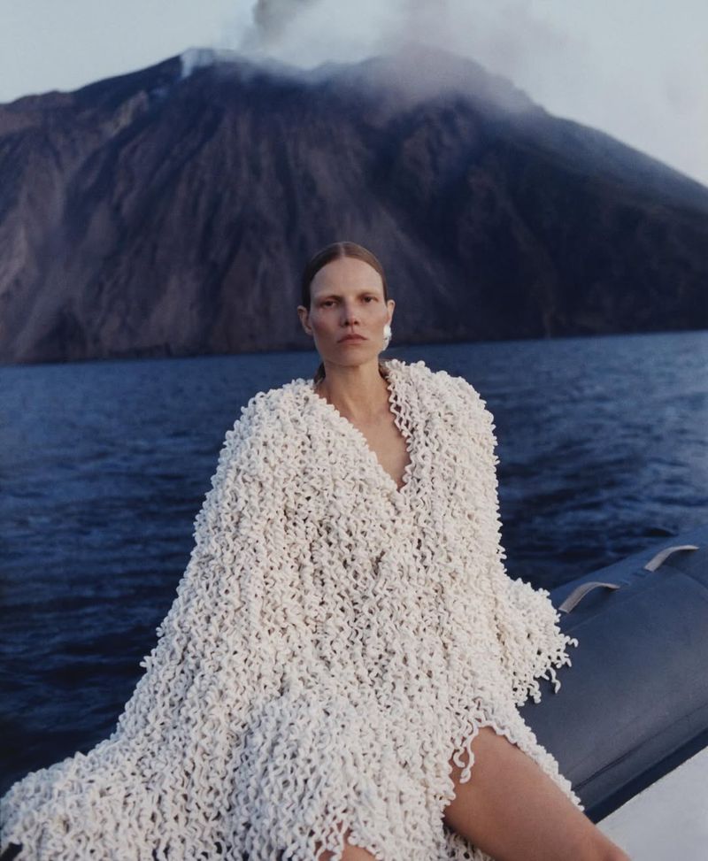 A fair-skinned adult woman is posing in a flowing white fringed coat in front of the ocean and a volcanic mountain covered in clouds.