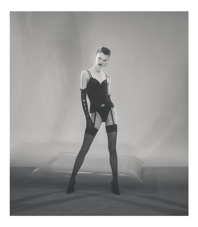 Monochrome fashion portrait of a woman in lingerie, stockings, and gloves standing in a studio setting with an abstract backdrop.