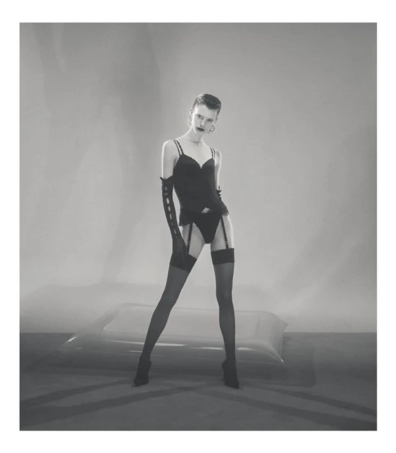 Monochrome fashion portrait of a woman in lingerie, stockings, and gloves standing in a studio setting with an abstract backdrop.