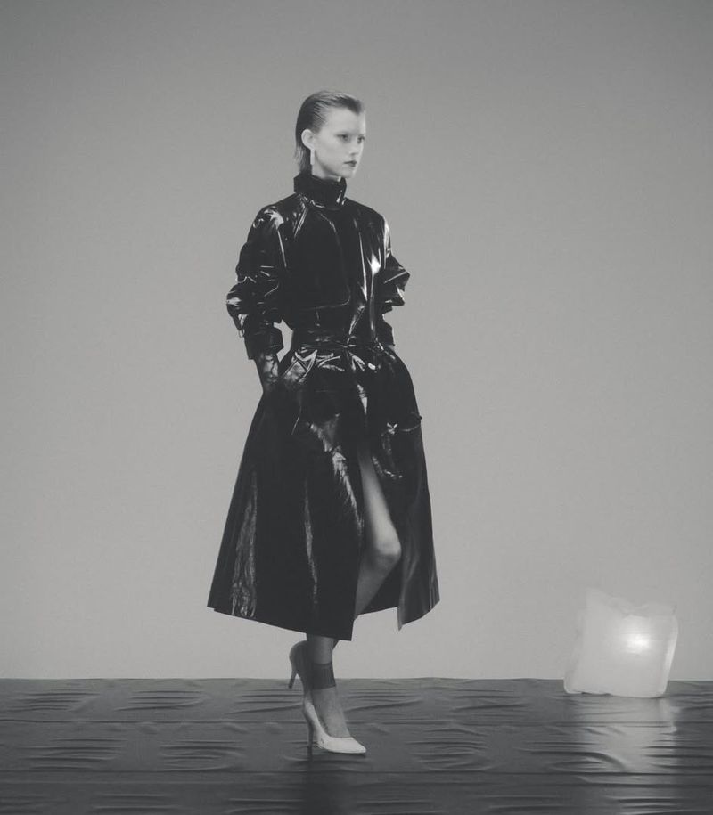 A lone adult female model poses in a shiny black vinyl coat with white high heels in a minimalist grey studio environment.
