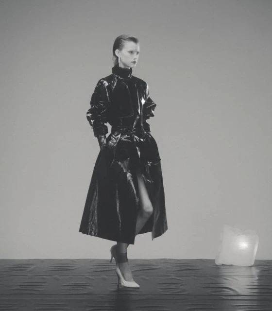 A lone adult female model poses in a shiny black vinyl coat with white high heels in a minimalist grey studio environment.