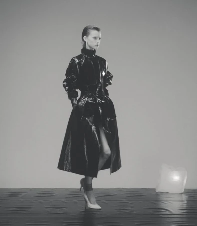 A lone adult female model poses in a shiny black vinyl coat with white high heels in a minimalist grey studio environment.