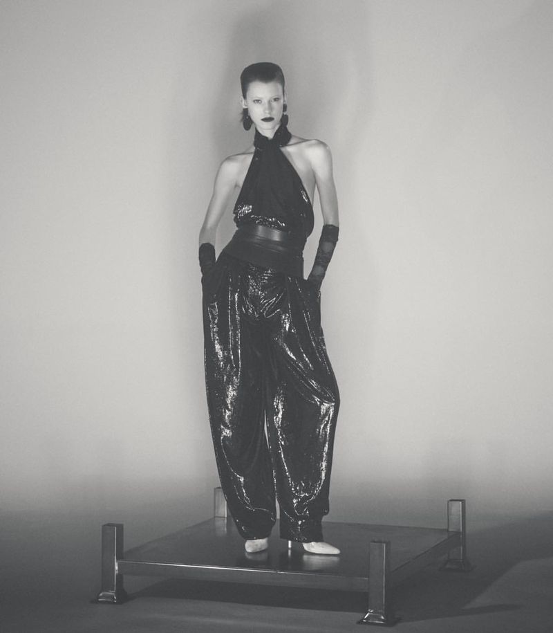 A tall, slender fashion model poses in a black halter-style top and flowing sequined pants on a metal platform against a neutral backdrop.