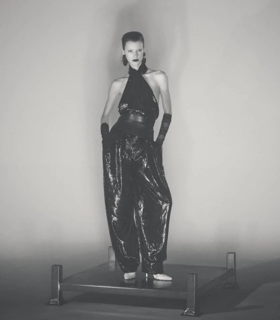 A tall, slender fashion model poses in a black halter-style top and flowing sequined pants on a metal platform against a neutral backdrop.