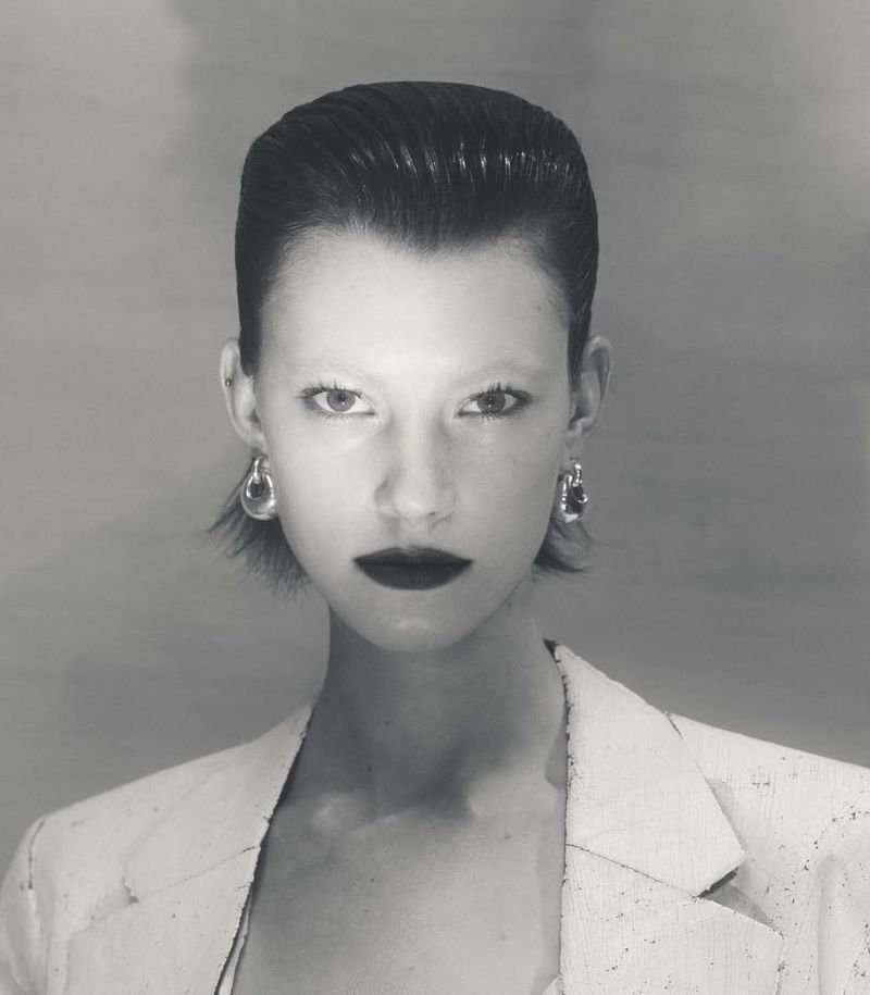 Portrait of a model with slicked-back dark hair, heavy dark lipstick, and hoop earrings wearing a white blazer against a neutral backdrop.