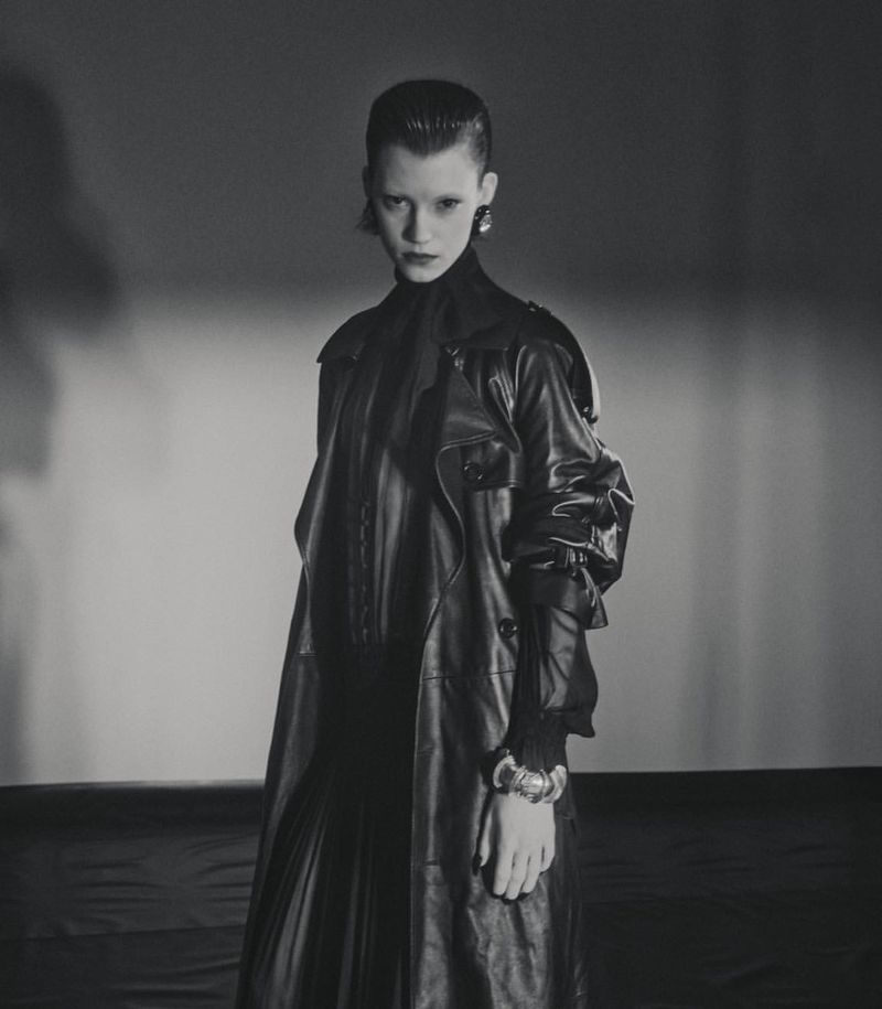 A woman with dark hair and an elegant earring poses in a black leather coat, looking at the camera against a gradient grey backdrop.