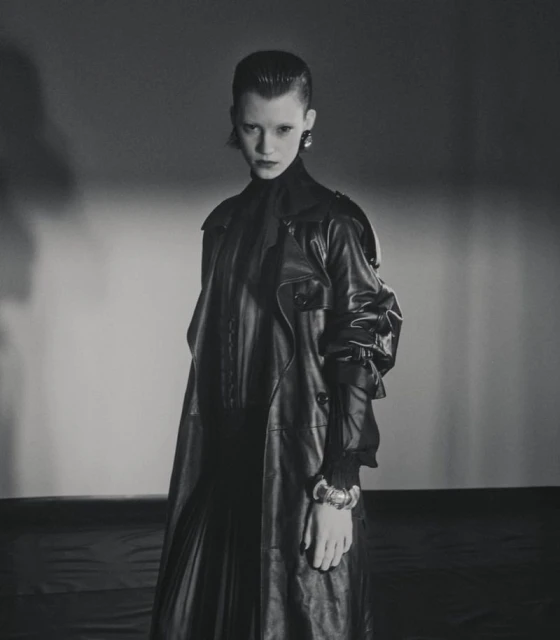 A woman with dark hair and an elegant earring poses in a black leather coat, looking at the camera against a gradient grey backdrop.