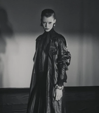 A woman with dark hair and an elegant earring poses in a black leather coat, looking at the camera against a gradient grey backdrop.