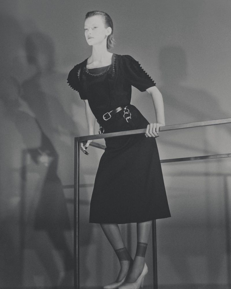 Fashion shot of a slim adult female model in a vintage black dress with tights and heels, posing against a gray backdrop, leaning on metal bars.