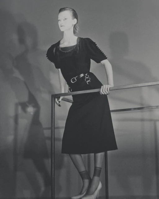 Fashion shot of a slim adult female model in a vintage black dress with tights and heels, posing against a gray backdrop, leaning on metal bars.