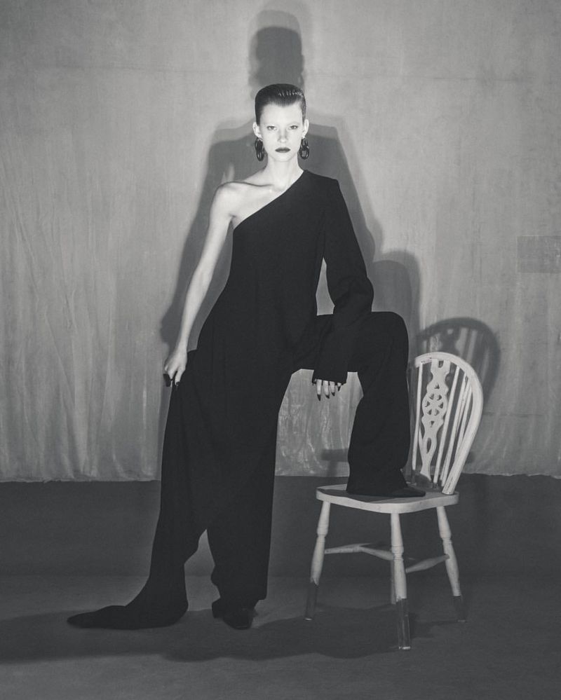 A slender woman poses in a one-shoulder black jumpsuit against a gray backdrop, seated on a painted chair with a high-fashion style.