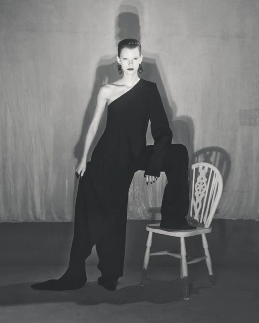A slender woman poses in a one-shoulder black jumpsuit against a gray backdrop, seated on a painted chair with a high-fashion style.