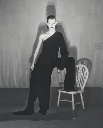 A slender woman poses in a one-shoulder black jumpsuit against a gray backdrop, seated on a painted chair with a high-fashion style.