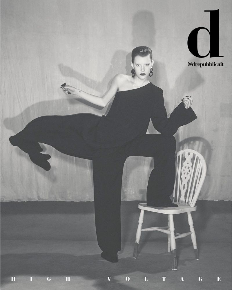 Monochrome fashion editorial shot of a slender woman in an oversized black suit posing with one foot on a white chair, studio setting.