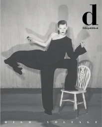 Model balancing on a white chair in a black one-shoulder top and trousers against a textured grey studio wall.