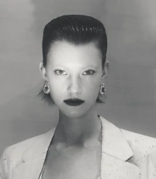 Portrait of a model with slicked-back dark hair, heavy dark lipstick, and hoop earrings wearing a white blazer against a neutral backdrop.