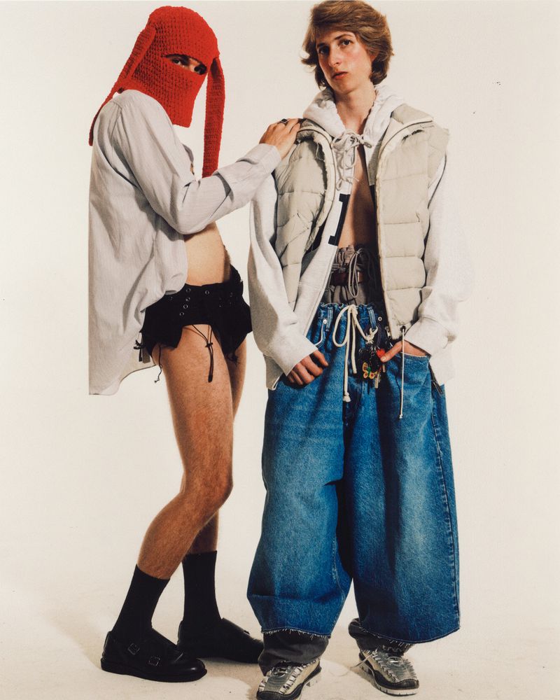 Two models strike a pose against a white backdrop. One wears a red balaclava, the other wears oversized jeans and a puffer vest.